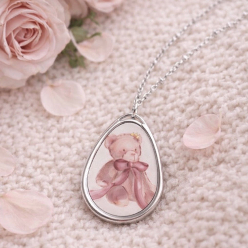 Teardrop-shaped pendant with a pink teddy bear design on a textured surface with pink roses.