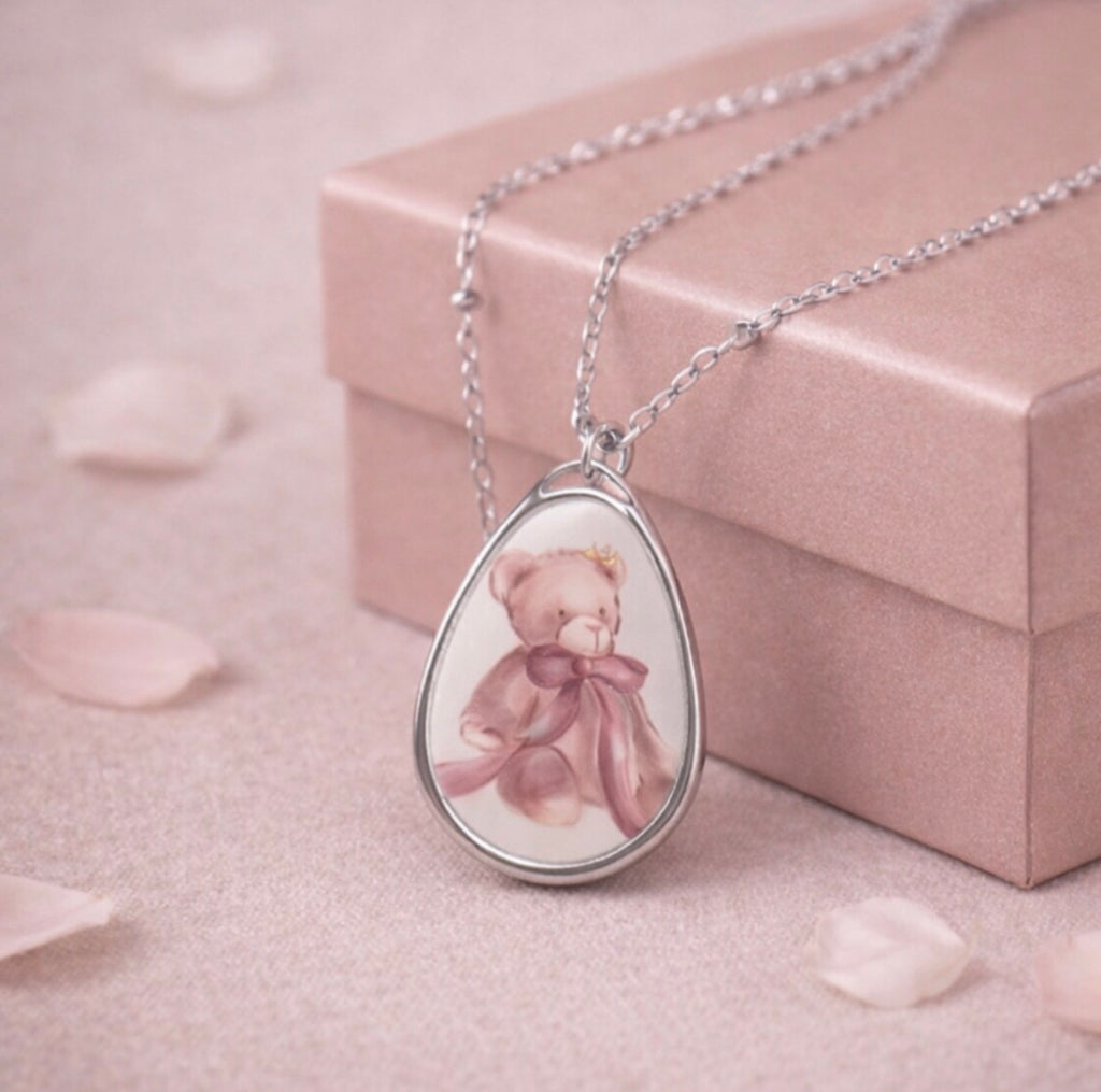 Teardrop-shaped pendant with a teddy bear design on a pink surface with a jewelry box in the background.