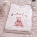 Folded white shirt with teddy bear design and 'Endless Love' text on a soft surface with a candle and cup.